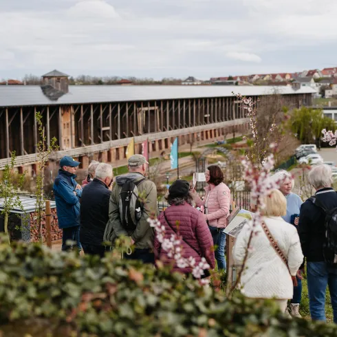 Eine Gruppe von Menschen steht in einem Garten und diskutiert. Im Hintergrund ist ein großes Gebäude zu sehen, und die Pflanzen beginnen zu blühen.