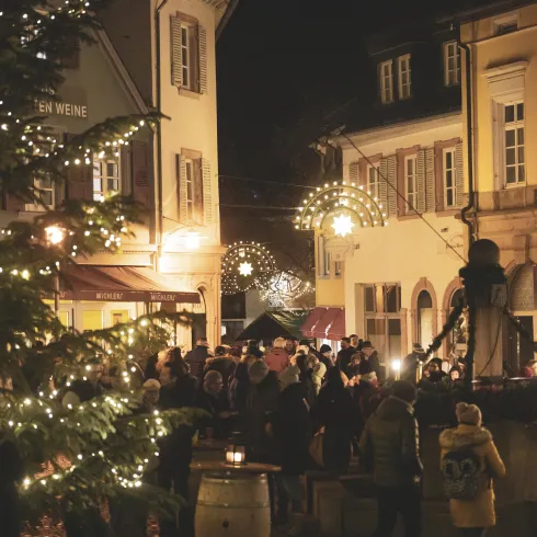 Ein festlicher Weihnachtsmarkt bei Nacht mit vielen Lichtern und fröhlichen Menschen. Im Vordergrund steht ein Weihnachtsbaum, umgeben von bunten Ständen.