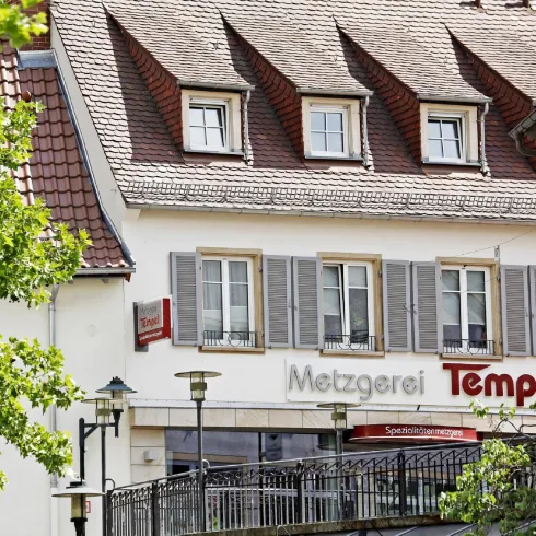 Ein Fachgeschäft mit dem Namen „Metzgerei Tempel“ befindet sich in einem charmanten Gebäude. Fenster mit Shutters und einladende Pflanzen vervollständigen das Bild.
