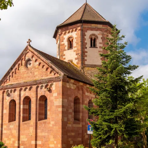 Eine historische Kirche aus rotem Sandstein steht auf einem gepflasterten Weg. Umgeben von Bäumen und charmanten Gebäuden ist die Szene ruhig und malerisch.