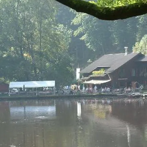 Ein idyllisches Gebäude am Ufer eines ruhigen Sees, umgeben von Bäumen. Menschen genießen die Natur und die friedliche Atmosphäre.