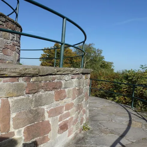 Eine steinerne Aussichtplattform mit einem Geländer. Umgeben von Bäumen und einem klaren blauen Himmel.