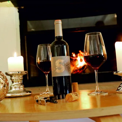 Ein Weinflasche steht auf einem Tisch neben zwei Weingläsern und Kerzen. Im Hintergrund flackert ein Kaminfeuer.