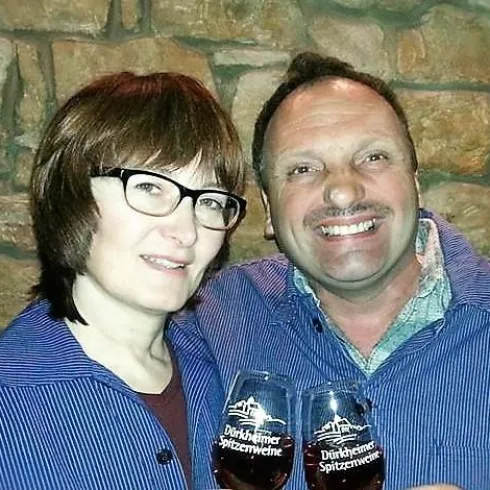 Ein Paar steht vor einer Steinwand und hält jeweils ein Glas in der Hand. Beide lächeln und tragen einheitliche, gestreifte Hemden.