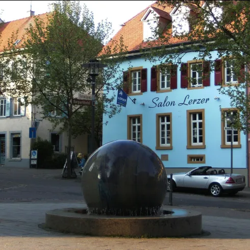Ein runder Brunnen mit sprudelndem Wasser steht im Vordergrund. Im Hintergrund sind bunte Gebäude und ein Parkplatz zu sehen.