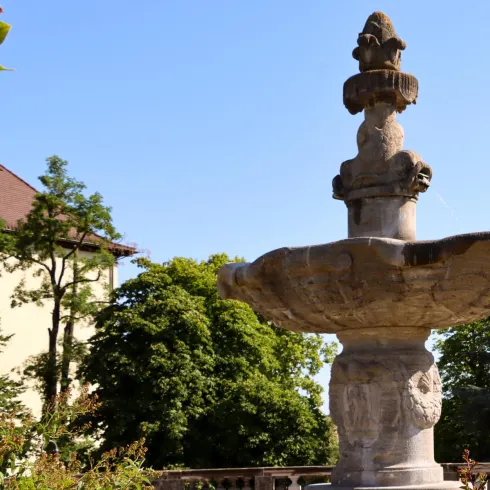 Ein schöner Brunnen in einem kreisförmigen Becken aus Stein. Das Wasser plätschert sanft, umgeben von einer ruhigen Landschaft.