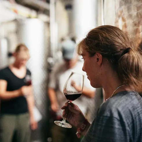 Eine Frau mit einem Glas Rotwein steht in einem Weinkeller. Im Hintergrund sind andere Personen und Weinfässer zu sehen.