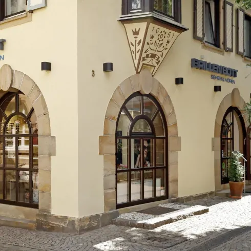 Ein charmantes Gebäude mit großen Bogenfenstern und einem Eingang. Die Fassade ist freundlich und einladend, umgeben von einer Pflanze in einem Blumentopf.