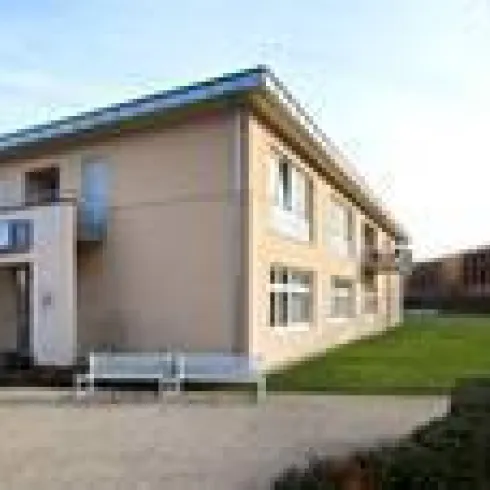 Ein modernes Gebäude mit hellbeiger Fassade und großen Fenstern. Im Vordergrund liegt eine gepflegte Grünfläche mit Sitzgelegenheiten.