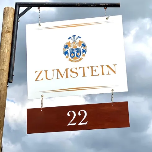 Ein Schild mit dem Namen "ZUMSTEIN" und der Nummer 22. Der Hintergrund ist bewölkt.