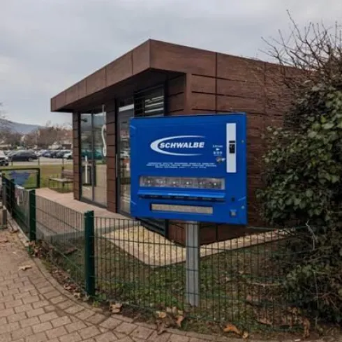 Ein blauer Automatständer mit dem Schriftzug „SCHWALBE“ steht an einem modernen Gebäude. Im Hintergrund sind Bäume und ein Gehweg zu sehen.