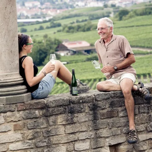 Zwei Menschen sitzen entspannt auf einer Mauer mit Blick auf Weinberge. Sie halten Gläser in der Hand und genießen die Aussicht.
