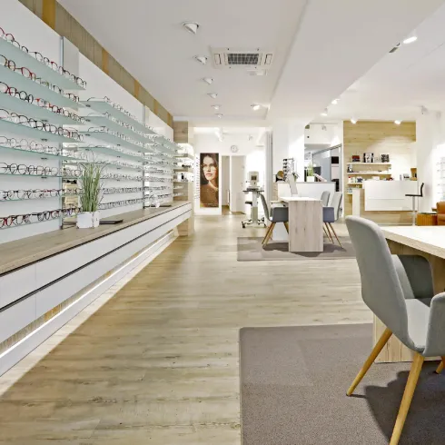 Ein modernes Geschäft für Brillen mit einer hellen, einladenden Atmosphäre. An der Wand sind zahlreiche Brillenrahmen ausgestellt, und es gibt gemütliche Sitzgelegenheiten.