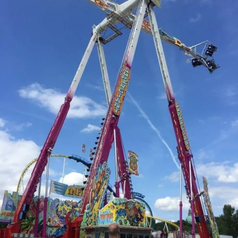 Ein großer Jahrmarktfahrgeschäft mit bunten Farben und einer beeindruckenden Struktur. Menschen stehen davor und beobachten die Attraktion unter einem klaren blauen Himmel.