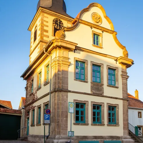 <p><p>Ein historisches Gebäude, das Leistadter Rathaus, mit einer markanten Fassade und einem schönen Dach. Das Haus ist im barocken Stil gehalten, die Fenster sind grün.</p></p>