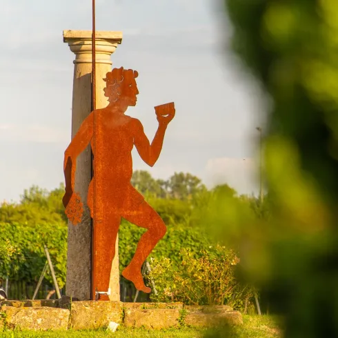 Eine rostige Skulptur eines tanzenden Mannes steht vor einer Säule. Im Hintergrund sind Weinreben und eine grüne Landschaft zu sehen.