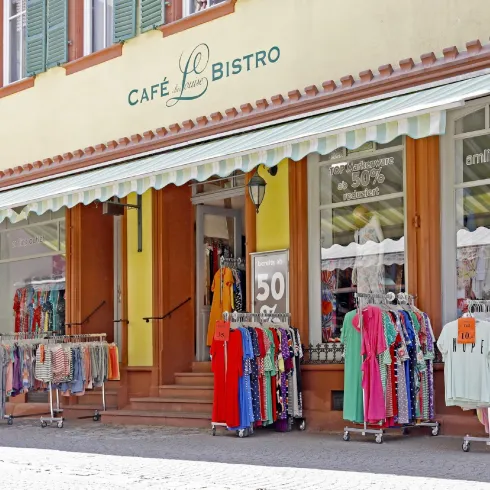 Ein charmantes Café mit Bistro und einer farbenfrohen Auswahl an Kleidung vor dem Eingang. Die Auslage bietet verschiedene Kleider auf Ständern zum Verkauf.