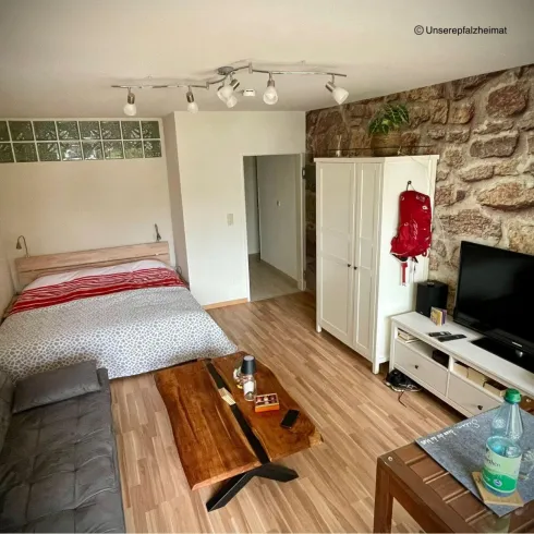 Ein modernes Zimmer mit einem Doppelbett, einer Couch und einem TV-Schrank. Die Wände sind elegant mit Steinen gestaltet, und die Einrichtung ist schlicht und einladend.