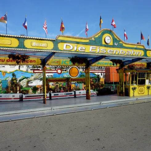 Ein bunter Bahnhofsbereich mit dem Schriftzug "Die Eisenbahn". Viele Flaggen schmücken die Umgebung und ein Freizeitpark-Feeling ist spürbar.