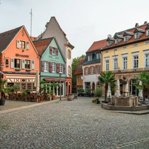 Ein malerischer Platz mit bunten Fachwerkhäusern und Restaurants. Die Umgebung ist mit Pflanzen und Blumen geschmückt und vermittelt eine gemütliche Atmosphäre.