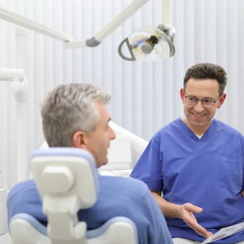 Ein Zahnarzt konsultiert einen Patienten in einer modernen Praxis. Beide tragen smiley Blicke und der Zahnarzt zeigt auf ein Blatt Papier.