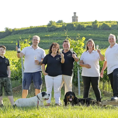 Eine Gruppe von sechs Personen steht in einem Weinberg und hält Gläser hoch. Zwei Hunde liegen am Boden, während sanfte Hügel im Hintergrund sichtbar sind.