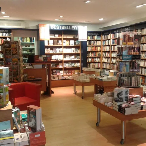 Ein gemütlicher Buchladen mit vielen Regalen voller Bücher. Im Vordergrund stehen Tische mit weiteren Büchern und einem roten Sessel.