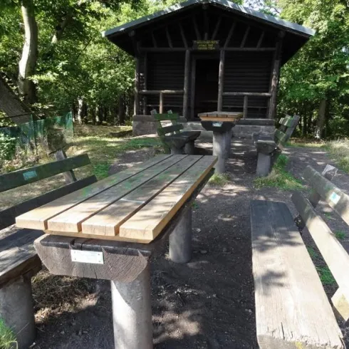 Eine gemütliche Sitzgruppe aus Holz mit Tisch und Bänken in einem Waldgebiet. Im Hintergrund steht eine kleine Hütten.