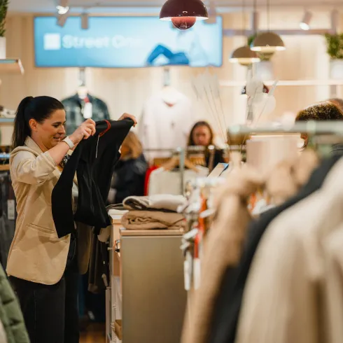 Eine Frau probiert Kleidung in einem modernen Bekleidungsgeschäft an. Im Hintergrund sind zahlreiche weitere Kunden und eine stilvolle Auswahl an Kleidung zu sehen.