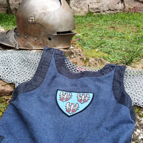 Ein mittelalterlicher Helm liegt neben einem blauen Wappenrock mit einem Emblem auf dem Boden.