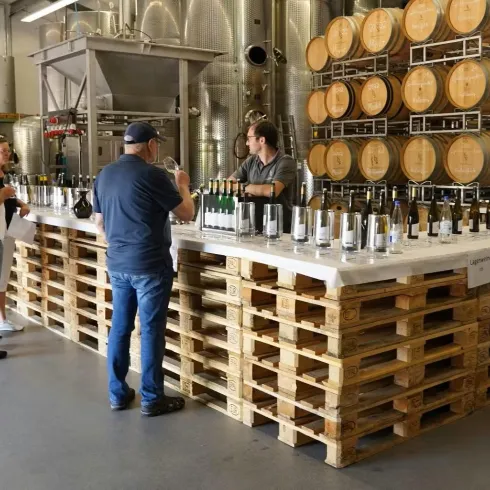 Eine Weinverkostung in einer Kellerwirtschaft. Menschen probieren verschiedene Weine an einem Tisch aus Holzpaletten mit Flaschen und Fässern im Hintergrund.