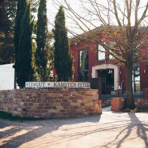 Ein Weingut mit dem Namen "Karsten Peter" und einer charmanten roten Fassade. Umgeben von hohen Zypressen und einem einladenden Eingangsbereich.