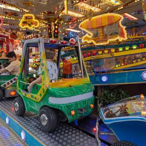 Ein bunter Vergnügungspark mit leuchtenden Lichtern und kinderfreundlichen Fahrgeschäften. Im Vordergrund sind kleine Autos zu sehen, in denen Kinder sitzen.