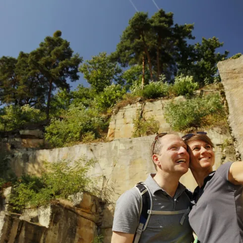 Ein Paar macht ein Selfie vor einer felsigen Kulisse mit Bäumen im Hintergrund. Der Himmel ist blau und die Sonne scheint.