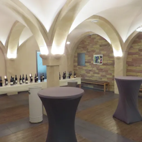 Ein Weinkeller mit eleganten Gipsbögen und Holzfußboden. Auf dem Tisch stehen viele Flaschen Wein, daneben gibt es Stehtische.