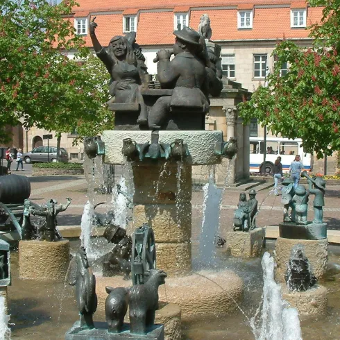 Ein schöner Brunnen mit skulpturalen Tierdarstellungen und sprudelndem Wasser. Im Hintergrund sind Blumen und Bäume zu sehen.