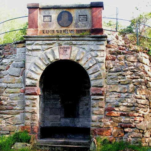 Ein historisches Gebäude aus Stein mit einem großen Rundbogen. Über dem Eingang befindet sich ein Schild mit der Aufschrift "Kaiser Wilhelms Höhe".