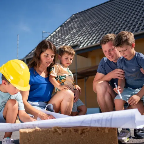 Eine Baustelle auf einer Wiese mit einem Bagger und mehreren Personen. Kinder spielen im Vordergrund, während Erwachsene darauf achten, dass alles sicher ist.