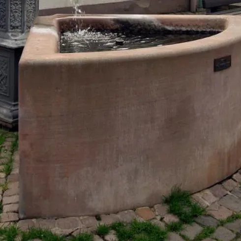Ein runder Brunnen aus Stein mit Wasser, das sprudelt. Er steht auf einem gepflasterten Boden und ist von grünem Gras umgeben.
