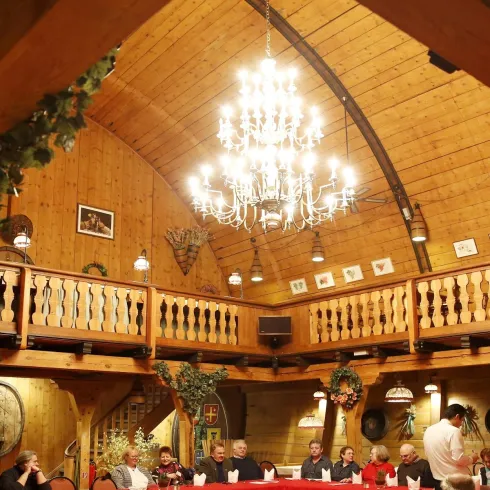 Ein gemütliches Restaurant mit Holzdekor und einer großen Decke. Viele Gäste sitzen an einem langen Tisch, während ein Kellner im Hintergrund bedient.