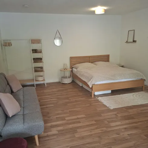 Ein helles Schlafzimmer mit einem großen Bett und einem gemütlichen Schlafsofa. Der Raum hat Holzfußboden und einfache, moderne Möbel.