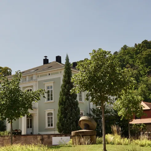 Ein elegantes Gebäude umgeben von Bäumen und Grünflächen. Im Hintergrund sind sanfte Hügel zu sehen.