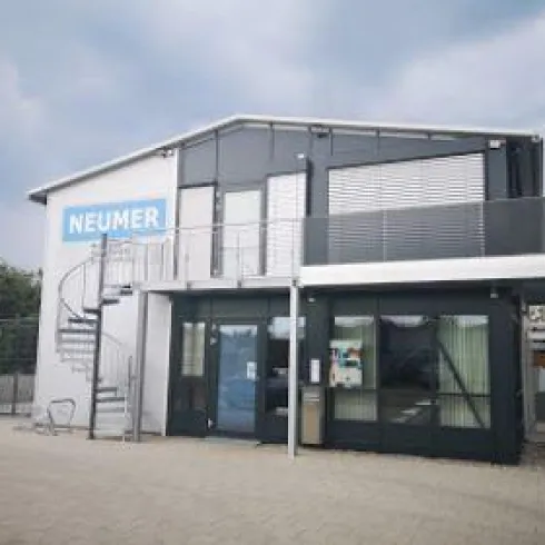 Ein modernes Gebäude mit einer geschwungenen Treppe und großen Fenstern. Die Fassade ist schwarz-weiß und trägt das Schild "NEUMER".