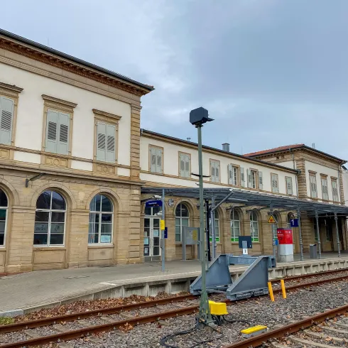 Ein Bahnhof mit historischem Gebäude und Gleisen. Der Himmel ist bewölkt und es gibt wenige Menschen in der Nähe.