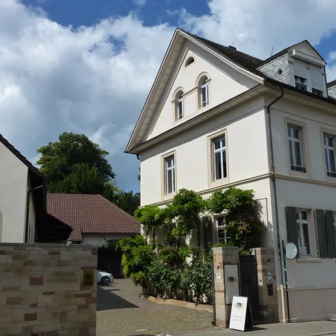 Ein schönes Gebäude mit einer hellen Fassade und grünen Pflanzen vor der Tür. Der Himmel ist teilweise bewölkt und die Umgebung wirkt ruhig.