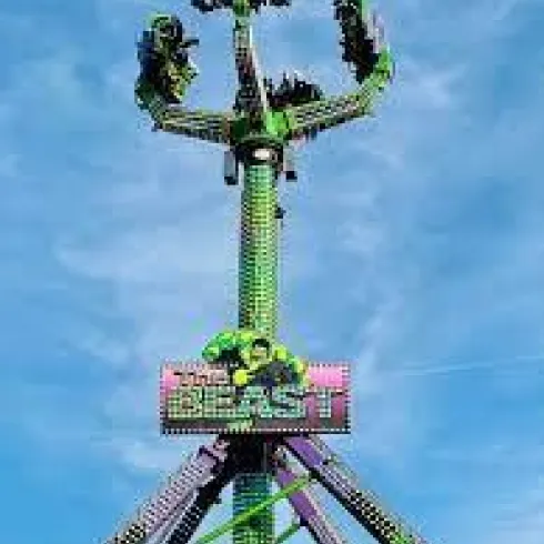 Ein hoher Fahrgeschäft mit grüner Beleuchtung, das die Fahrgäste in die Höhe und Drehung bringt. Der Himmel ist blau und klar.