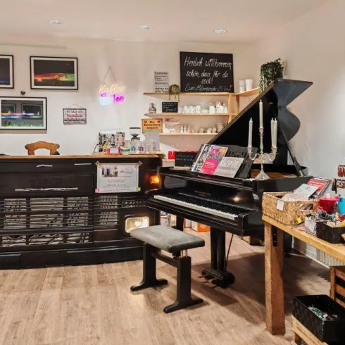 Ein gemütlicher Raum mit einem Klavier und einem Empfangstresen. Wanddekorationen und Geschenktische schaffen eine einladende Atmosphäre.