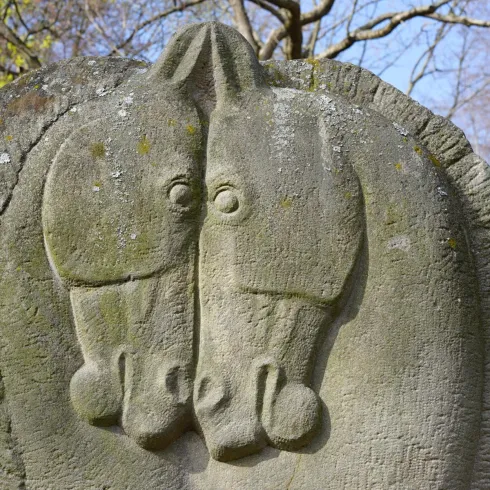 Eine Stein Skulptur mit zwei Pferdeköpfen. Die Statue steht in einem Park mit Bäumen im Hintergrund.