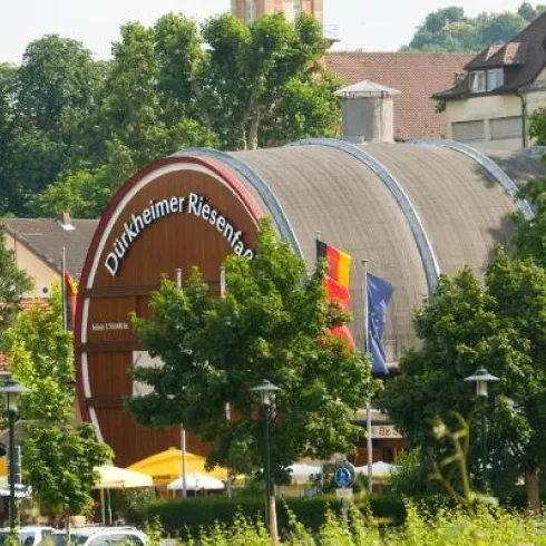 Ein großes, rundes Gebäude mit dem Schriftzug "Durlheimer Riesenfass" ist von Bäumen und Grünflächen umgeben. Im Vordergrund sind einige Tische und Stühle im Freien zu sehen.