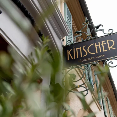 Ein Schild der Goldschmiede "Kinscher" ist an einem Gebäude befestigt. Im Vordergrund sind grüne Pflanzen sichtbar.